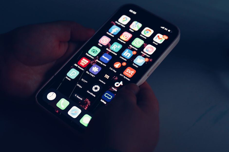 Close-up of hands holding a smartphone displaying various app icons in a dimly lit room.