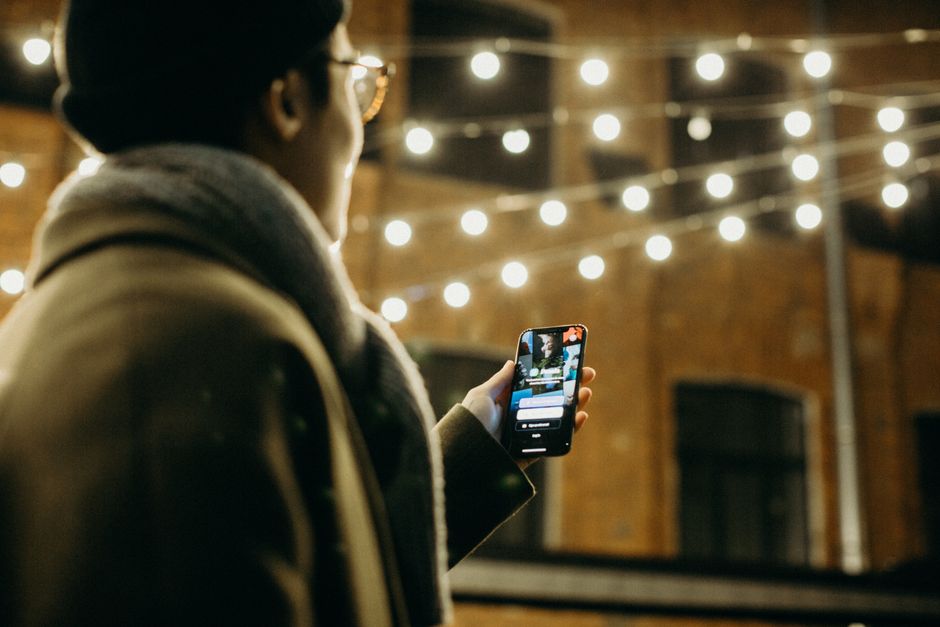 A person using a smartphone outdoors at night, illuminated by warm string lights.