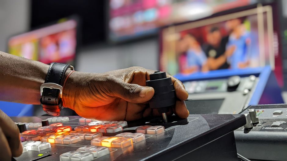 A hand operates a broadcast control mixer in a television studio setting.