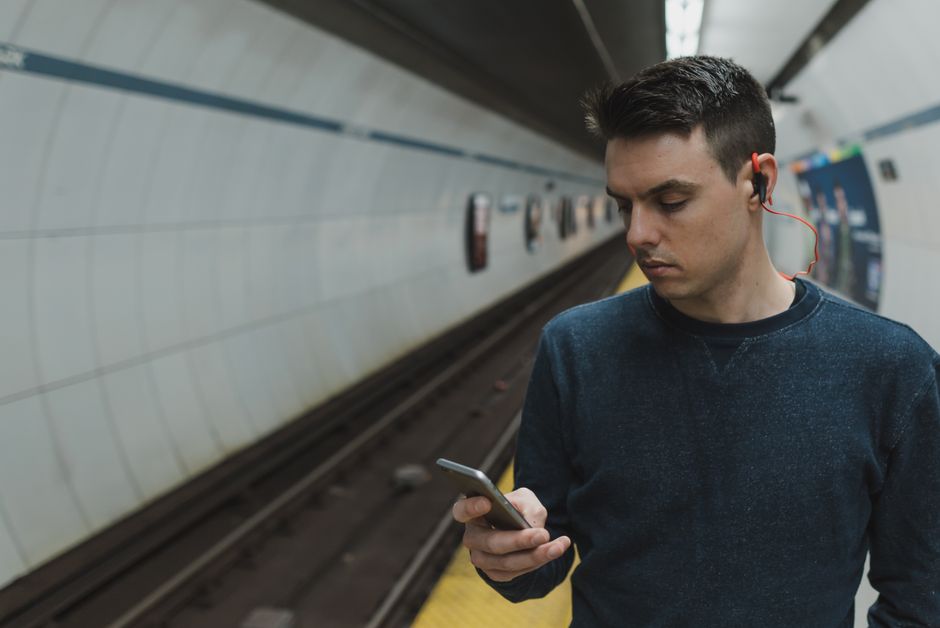 Man using a smartphone with earphones in a subway station, depicting modern urban lifestyle.